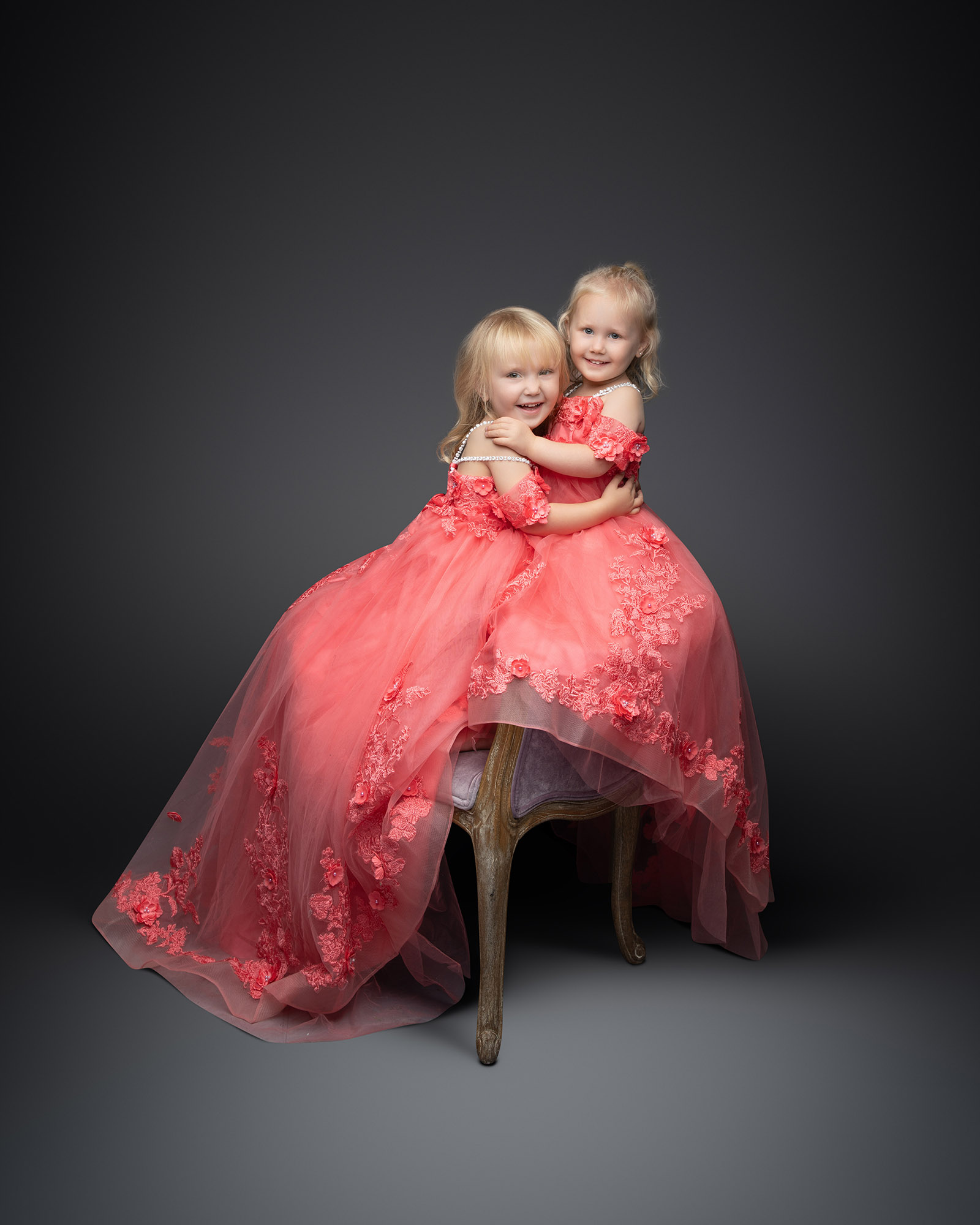 Two young sisters hugging while wearing elegant, floor-length pink floral dresses.