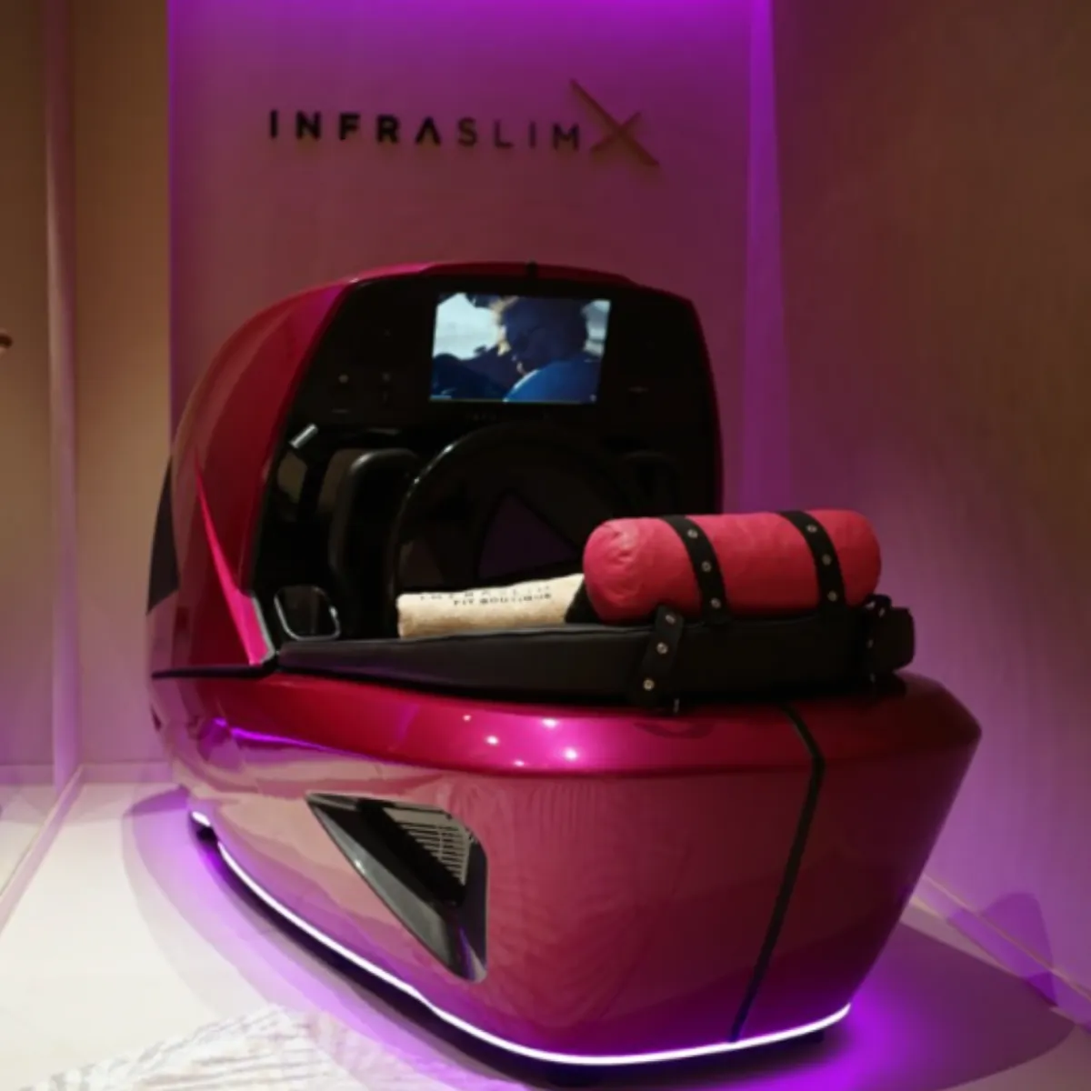 InfraslimX machine in a vibrant pink design with glowing LED lights. The machine features a screen at the back and a rolled towel on the seat, creating a relaxing and modern atmosphere in the treatment room.