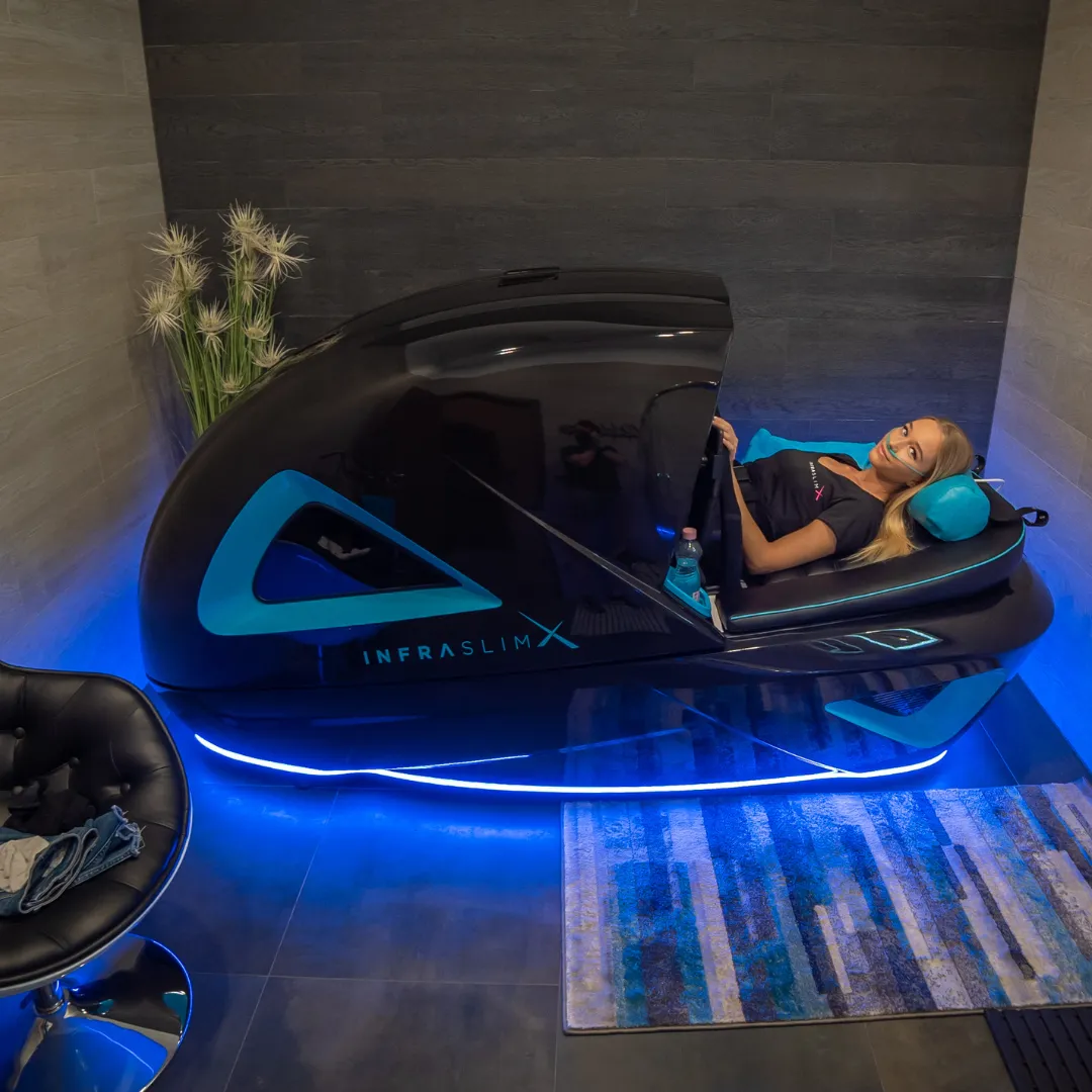 A woman relaxing inside an InfraslimX machine, illuminated by blue LED lights. The sleek black design with turquoise accents is visible as she lies back with a headrest, in a modern, spa-like setting.