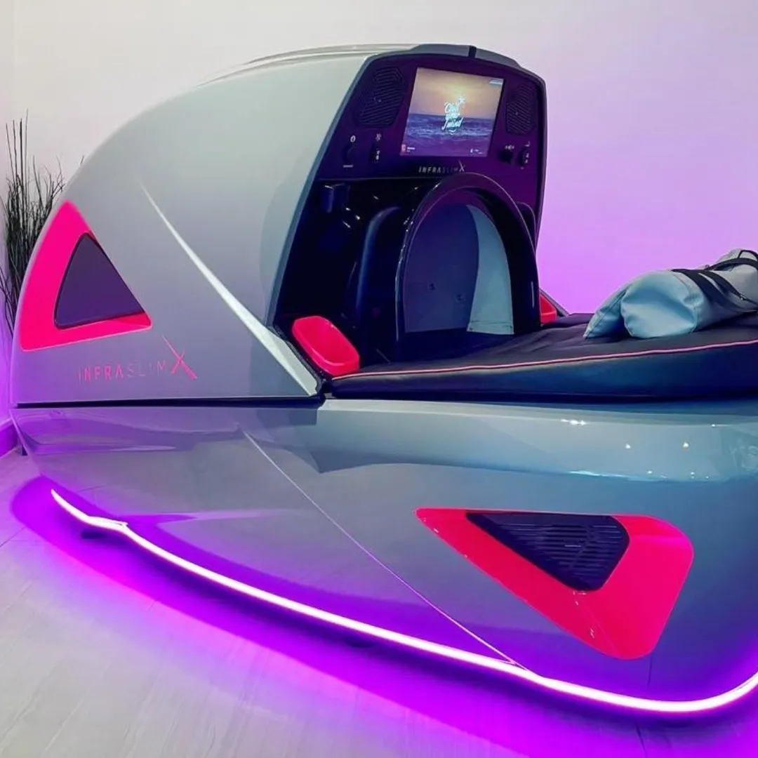 InfraslimX machine in sleek gray with vibrant pink accents and LED lighting. The machine is open, showing a comfortable seat with pillows, ready for a session. A screen is visible at the front, displaying calming content.