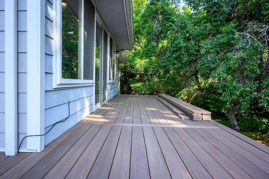 Seamless brown composite decking installed beside a white-sided house, blending with lush greenery for a tranquil outdoor living space.