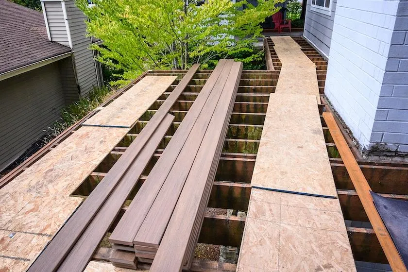 Residential deck construction in progress with composite decking material and pressure-treated wooden frame against a backdrop of lush greenery.