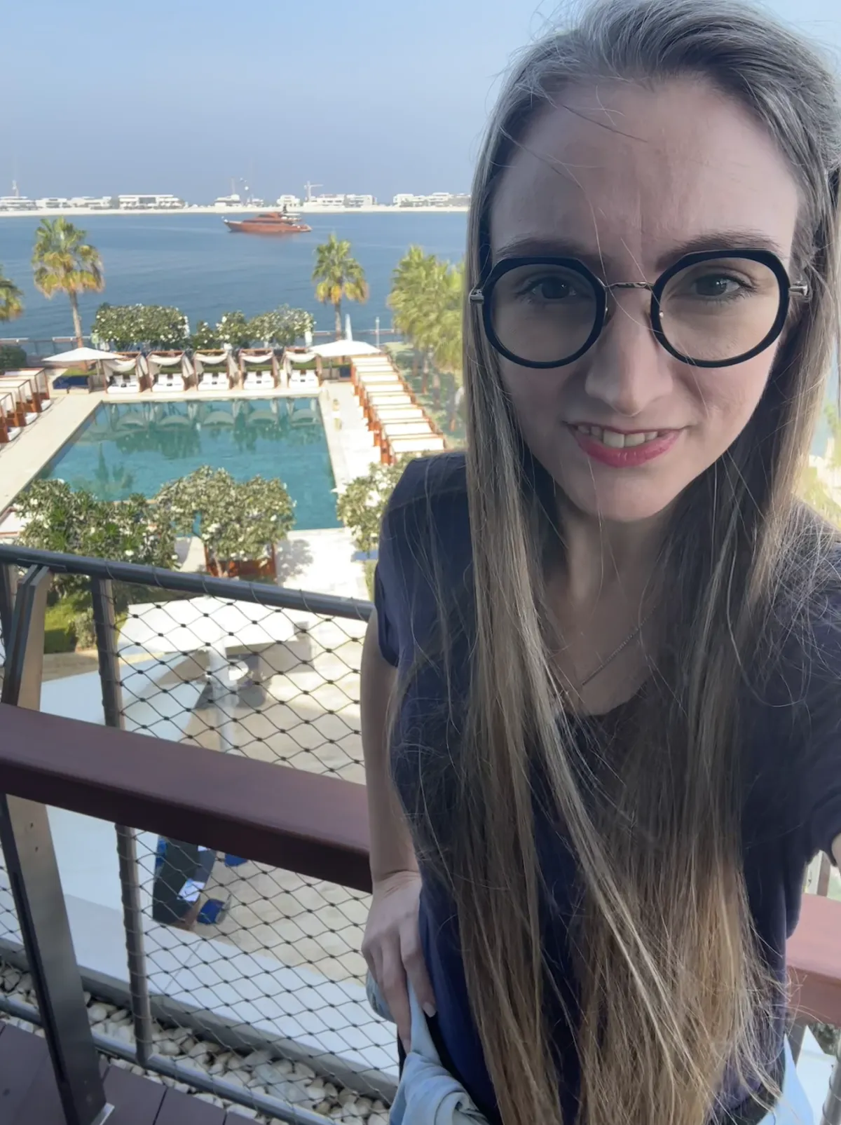 Martina Kikic, with long hair and glasses smiling on a balcony overlooking a luxury pool and waterfront view with palm trees and a docked ship in the background. The scene suggests freedom, travel, or lifestyle change, possibly tied to success on LinkedIn or with the Launchpad OS.