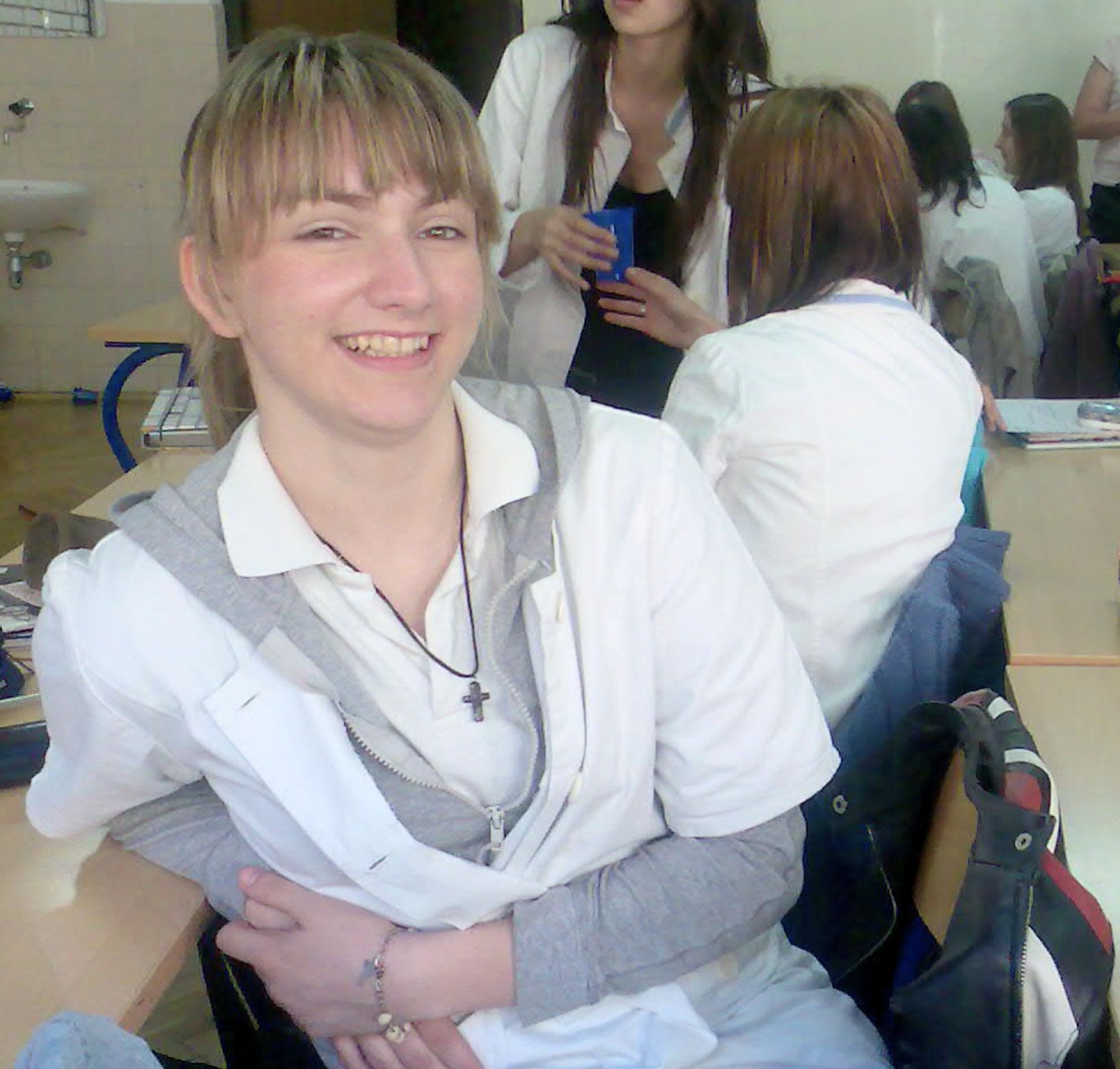 Young woman in a white lab coat smiling while seated in a classroom with other students in similar coats. She wears a cross necklace and leans slightly forward, with a relaxed and cheerful expression, suggesting a medical school environment.