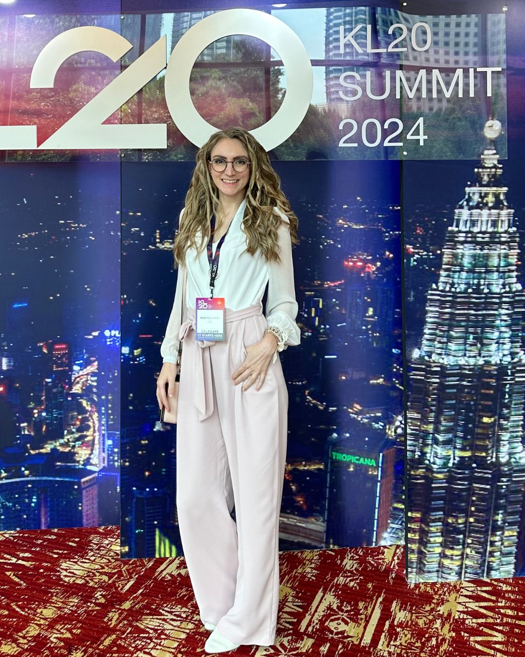 Martina Kikic in business attire stands smiling in front of a KL20 Summit 2024 backdrop featuring the Kuala Lumpur skyline. She wears a white blouse, pink wide-leg trousers, and a summit badge, conveying professional growth and thought leadership, potentially tied to success with LinkedIn Launchpad OS.