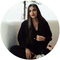 Confident young woman with long dark hair and blonde highlights, sitting casually in a stylish modern space. Personal brand photo for LinkedIn or professional bio.