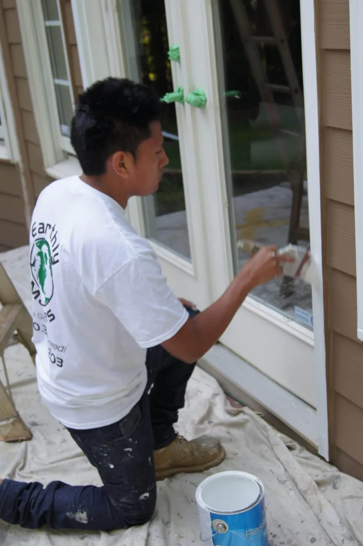 A painter kneels to apply a precise coat of white paint to the trim of an exterior glass door.