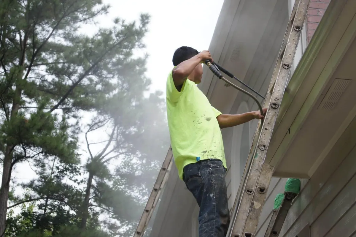 A close-up of a painter using a traditional block brush to hand-apply a coat of white limewash to a red brick home.