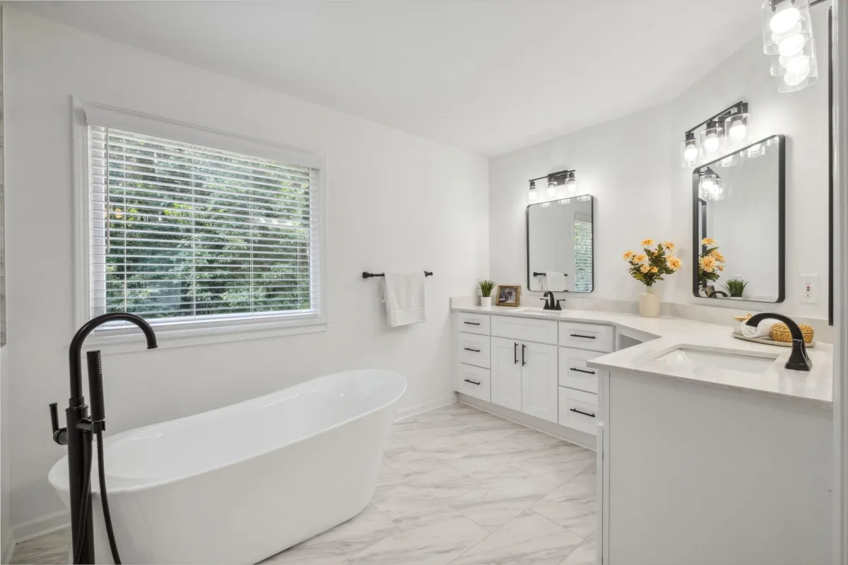 A clean and bright master bathroom with a double vanity, large mirrors, and a freestanding bathtub by the window.
