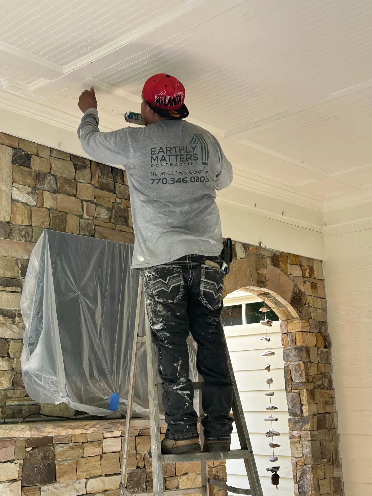 Painter in a red hat applying finish to an interior ceiling above a stone wall.