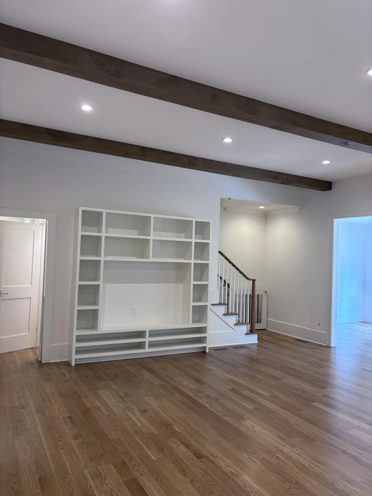 A spacious living room with freshly painted white walls, large custom built-in shelving, and dark wood ceiling beams.