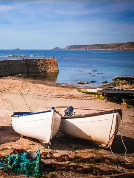 Two white fishing boats resting on the sand in the historic harbour of Sennen Cove, Cornwall, with the calm blue sea in the background
