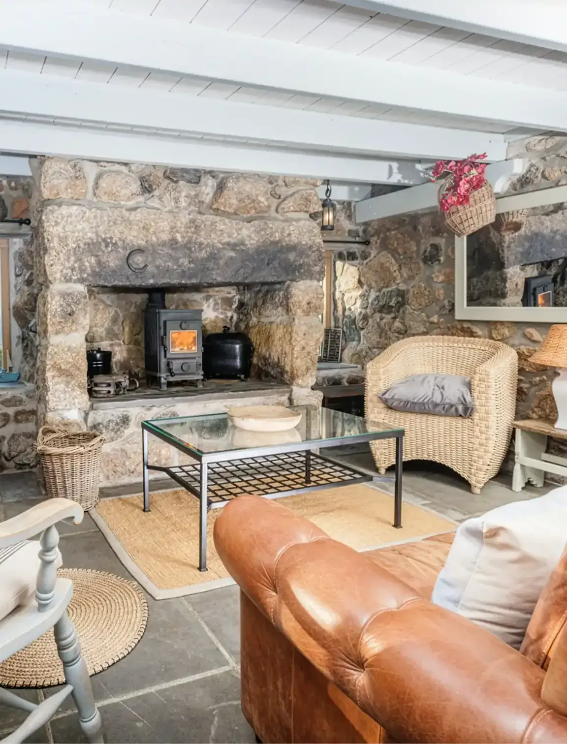 Cosy living room in a traditional Cornish stone cottage featuring a large granite inglenook fireplace with a lit wood-burning stove, leather sofa, and wicker armchairs.