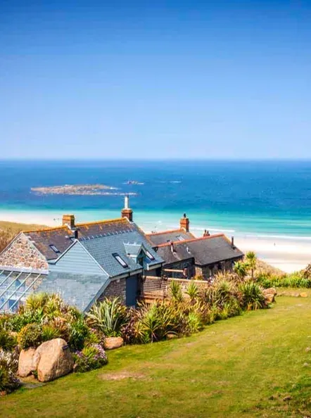 Aerial view of luxury holiday cottages nestled on a green cliffside overlooking the turquoise sea and white sands of Sennen Cove, Cornwall.