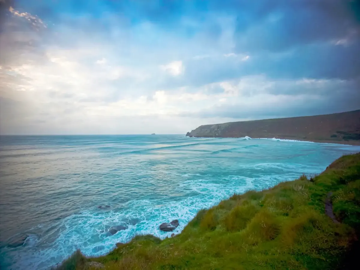 Dramatic view of rolling turquoise Atlantic waves meeting the rugged Cornish coastline near Sennen, under a dramatic cloudy sky.