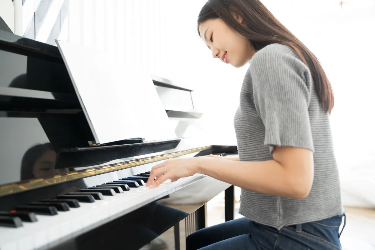An adult, dressed in a colorful long sleeve and eyeglasses, sits attentively at a sleek digital keyboard, their fingers moving gracefully over the keys. The soft glow of a tilt light bathes the scene, casting an inviting, warm ambiance around them. 