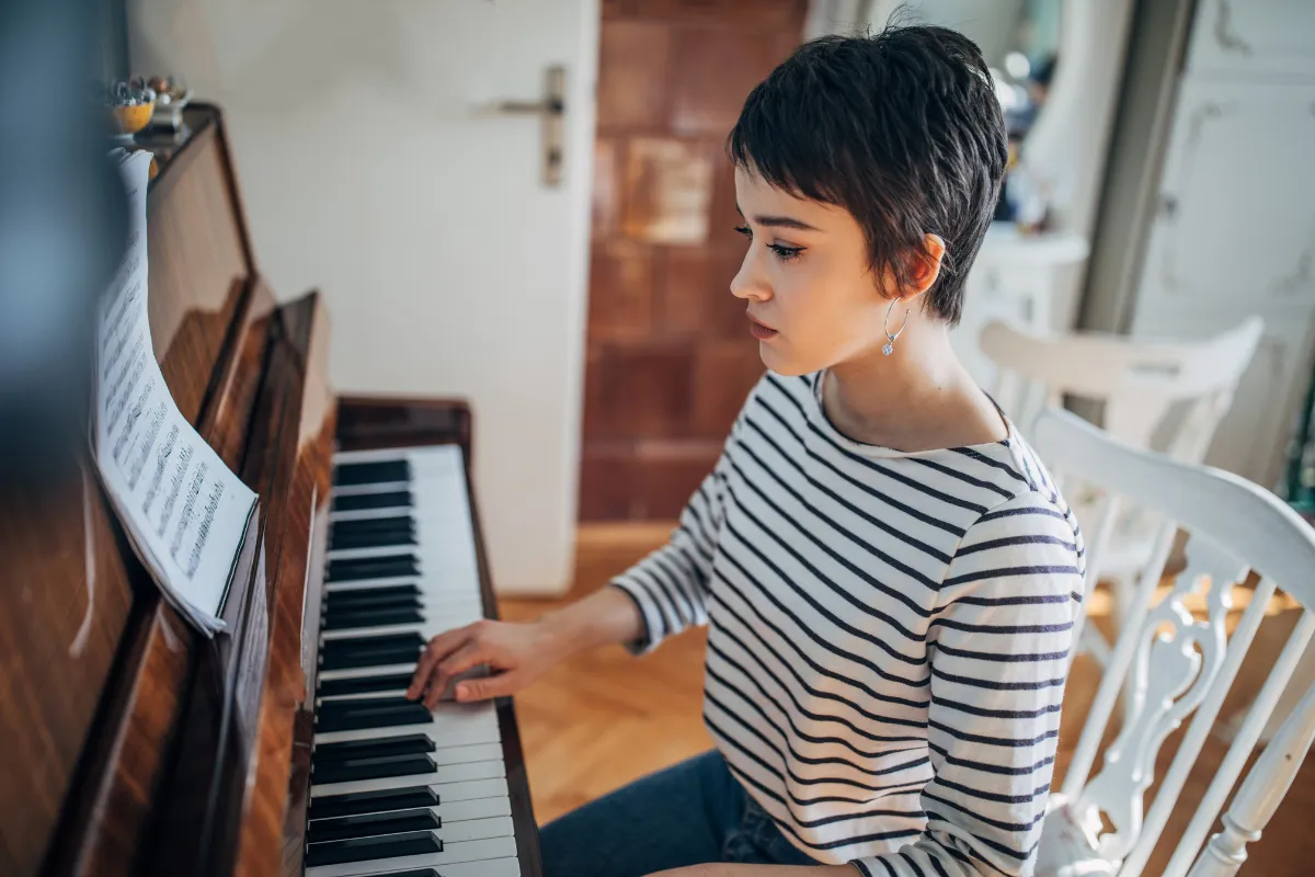 An adult, dressed in a colorful long sleeve and eyeglasses, sits attentively at a sleek digital keyboard, their fingers moving gracefully over the keys. The soft glow of a tilt light bathes the scene, casting an inviting, warm ambiance around them. 
