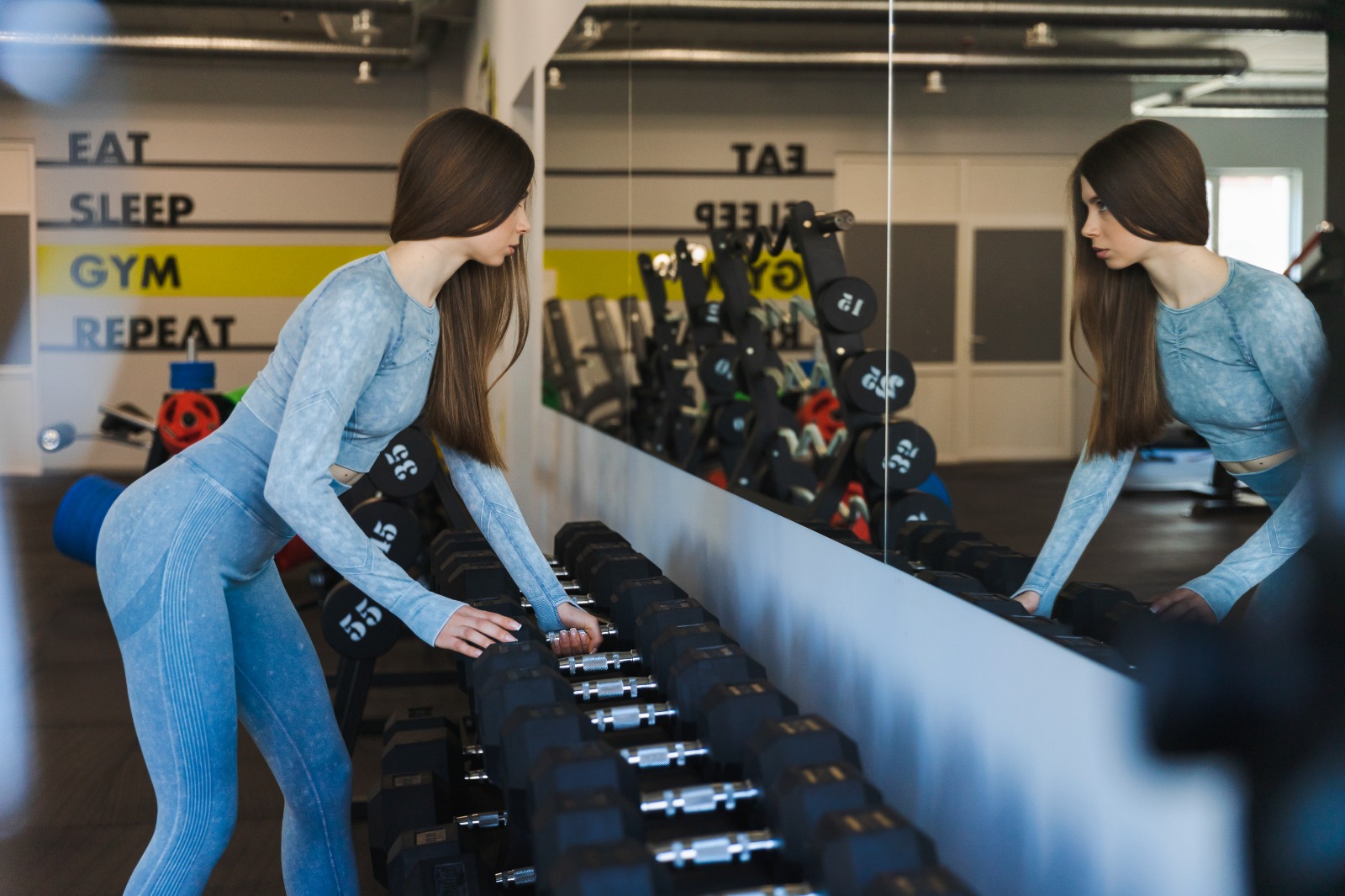 Fitness woman in blue workout gear selecting a dumbbell from a weight rack in a commercial gym, using the long wall-mounted gym mirror to check her form and reflection in the free weight area | Creoglass