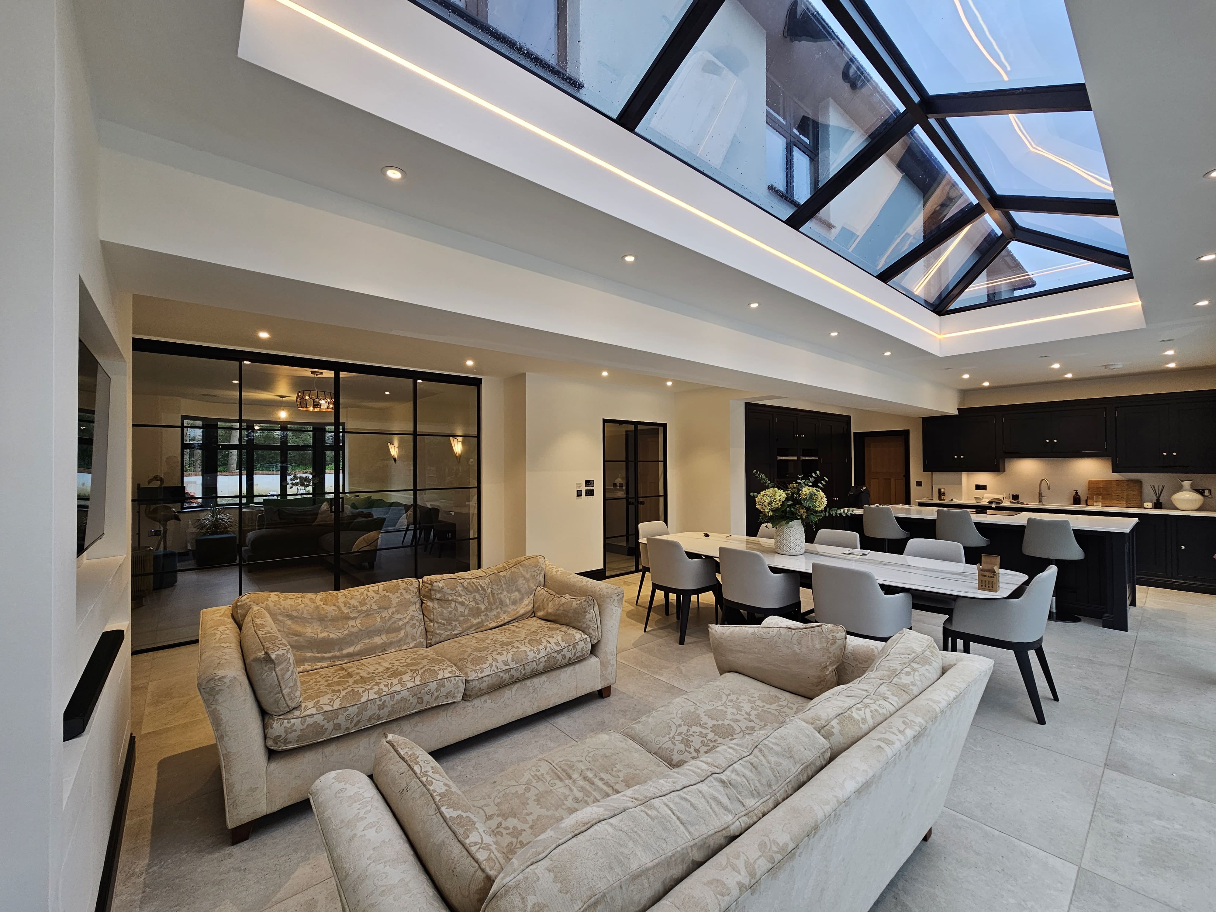 Wide black-framed glass sliding partition separating a living room and a modern kitchen under a glass roof. | Creoglass