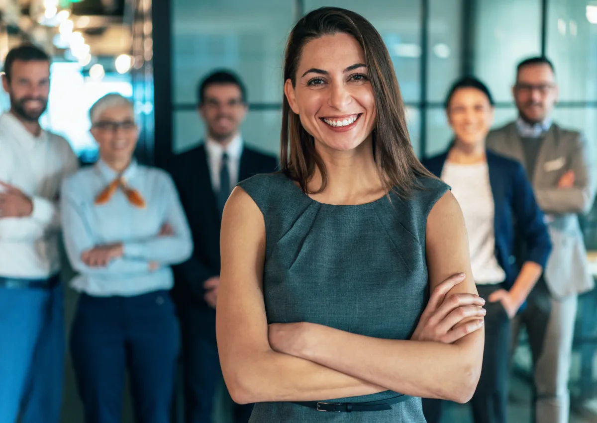 Mulher executiva sorrindo com braços cruzados e equipe de negócios ao fundo em ambiente corporativo