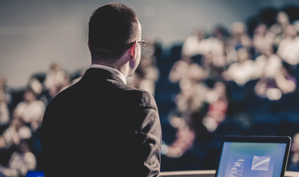 Palestrante de costas no palco observando plateia e audiência lotada em conferência corporativa