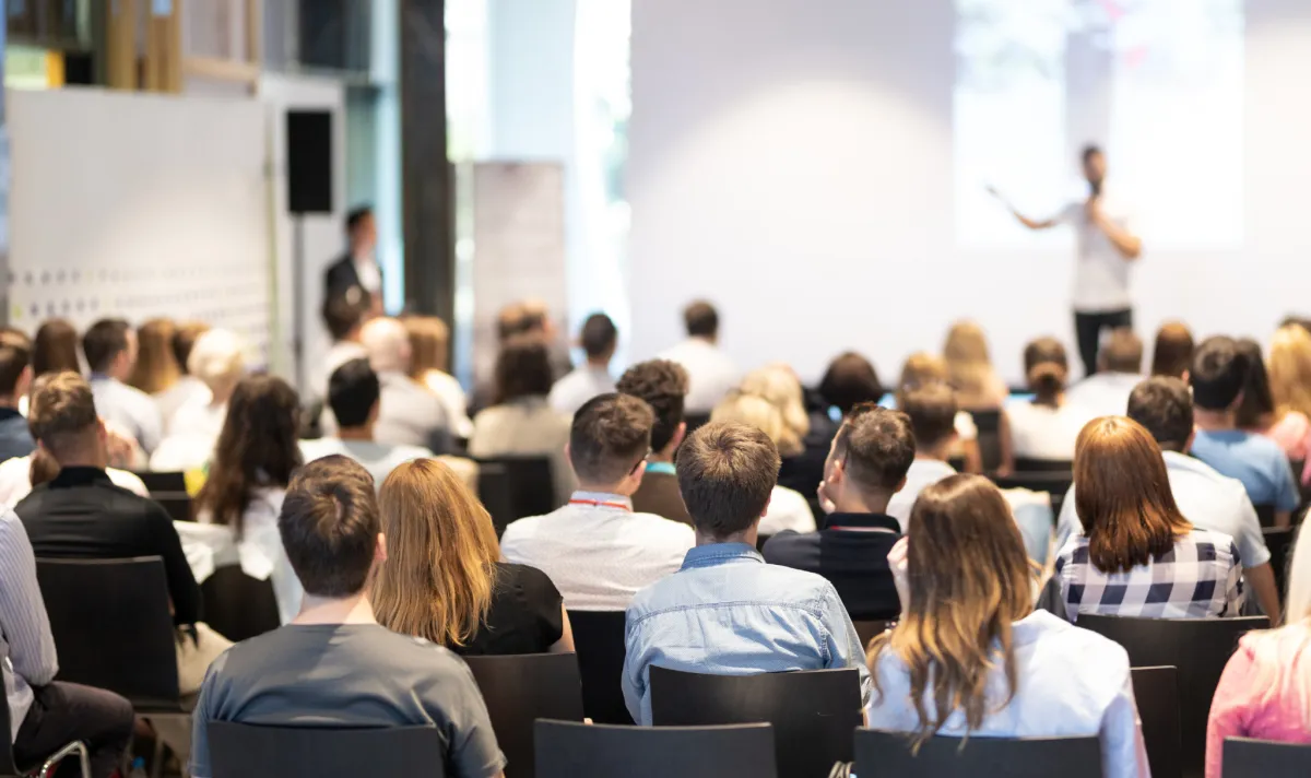 Plateia sentada em auditório assistindo palestrante no palco durante conferência ou evento corporativo