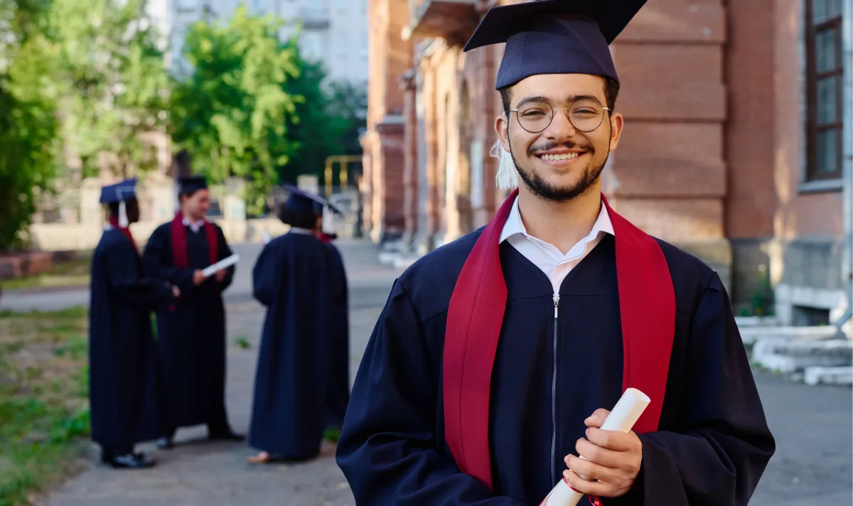 Estudante formando sorrindo de bata e capelo segurando diploma em frente ao campus universitário