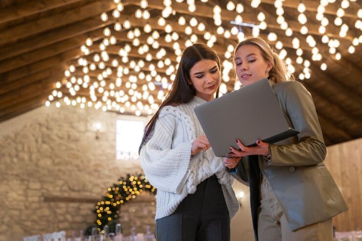 Duas organizadoras de eventos analisando projeto no notebook em salão decorado com luzes