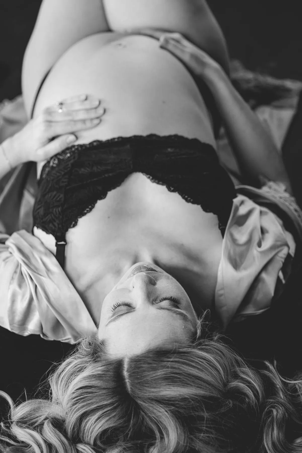 Grand Rapids maternity boudoir photography, pregnant woman laying on her back wearing a black bralet; hands on her bare pregnant belly.