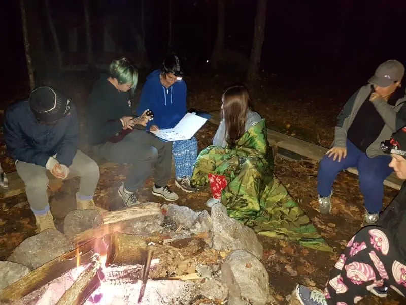 Students gathered around a campfire at The Ark, singing and playing music during an outdoor education program in Denholm, Quebec