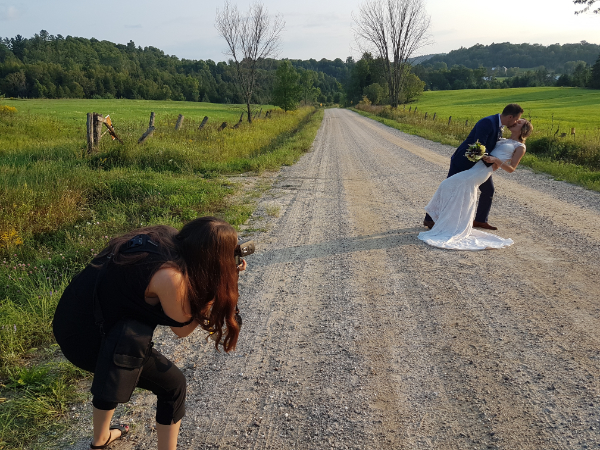 Wedding couple posing playfully on a gravel road while a photographer captures the moment