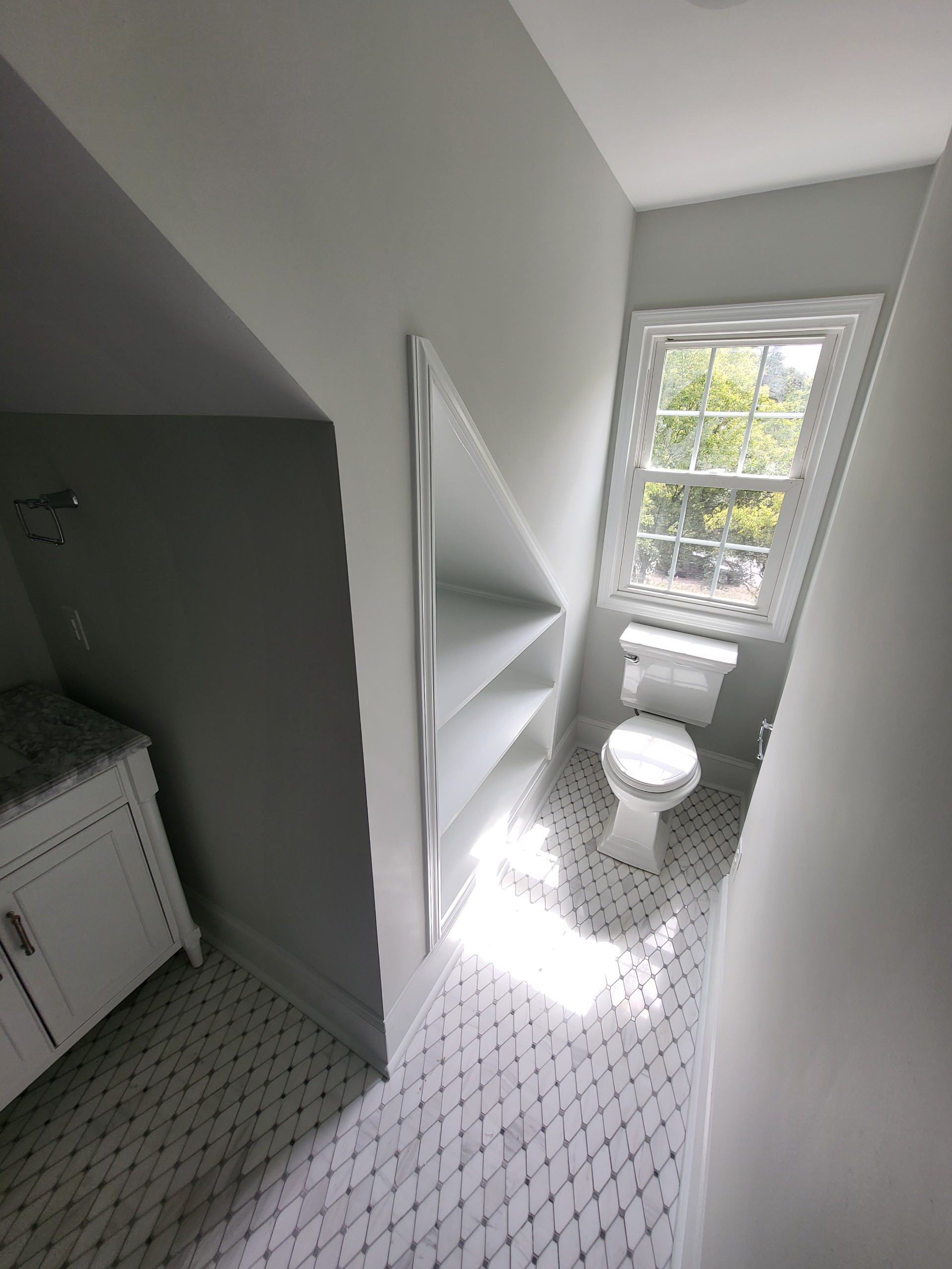 Small modern bathroom under staircase with built-in shelving, white toilet, marble tile floor, and window bringing in natural light.