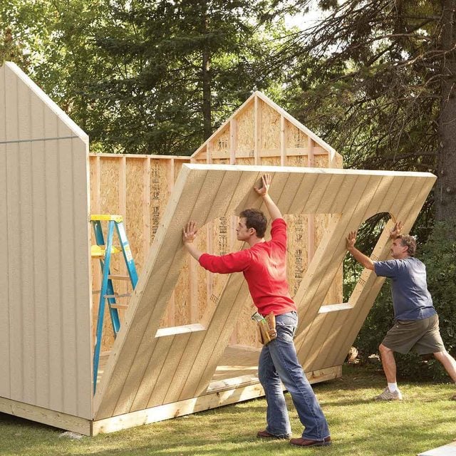 Workers installing a wall panel during backyard storage shed construction, outdoor wooden shed build.