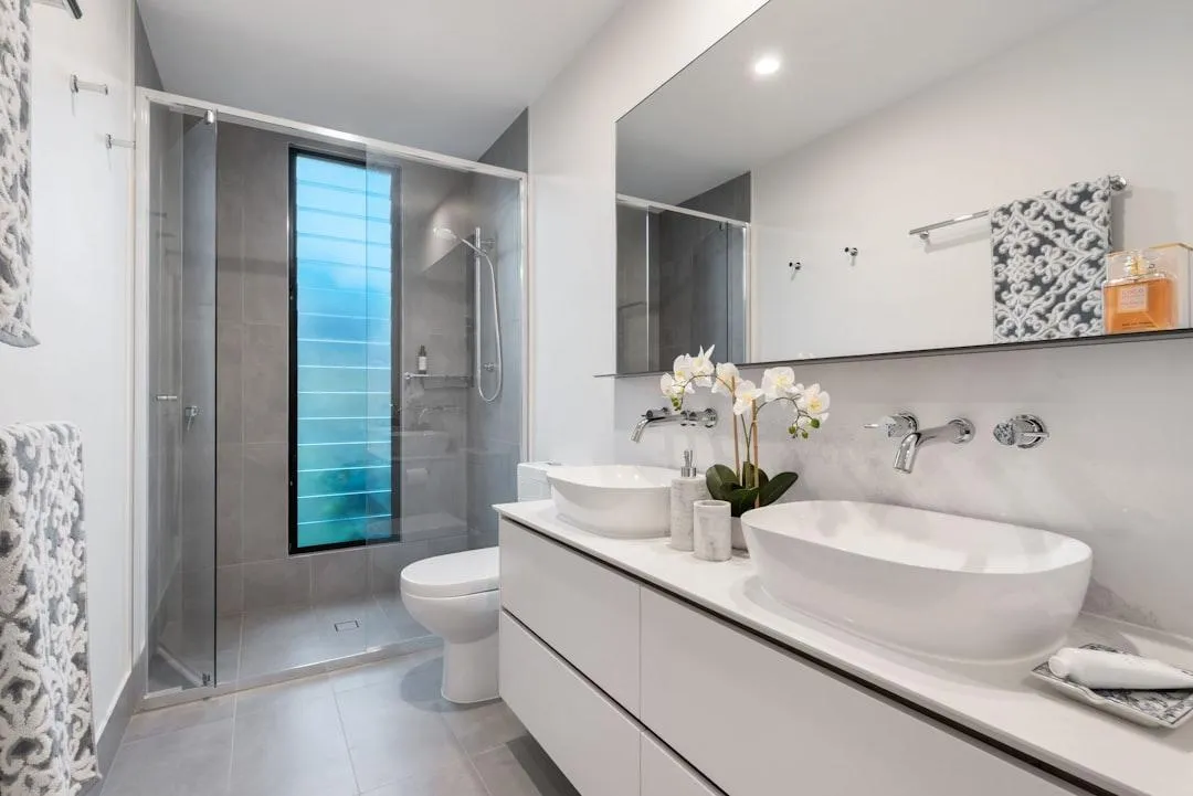 Modern minimalist bathroom with glass walk-in shower, double vessel sinks on a floating vanity, gray tile flooring, large mirror, and contemporary fixtures.