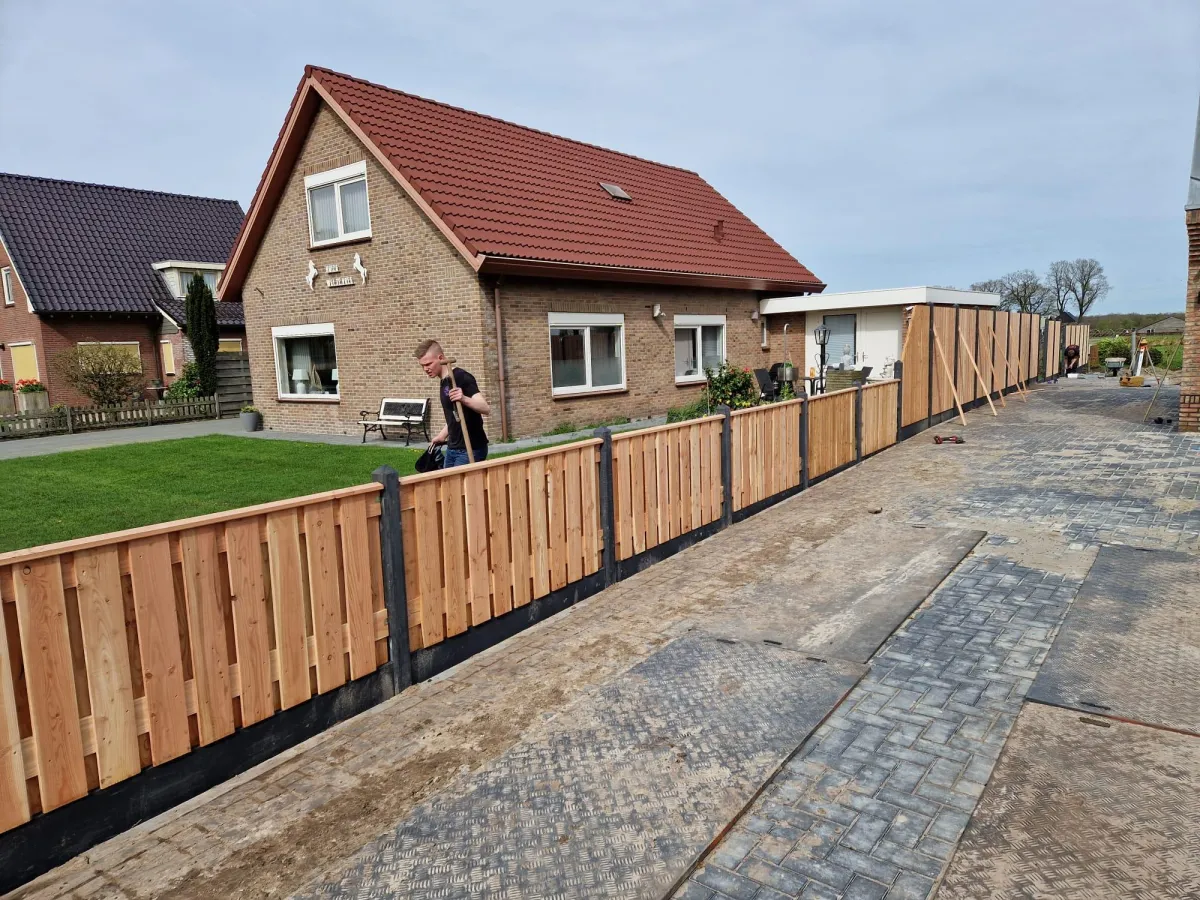 Twee ervaren vakmensen van Mulder's Multi Klus Service in werkkleding, lachend en geconcentreerd bezig met het plaatsen van een nieuwe schutting in een tuin. Gereedschap en materialen liggen netjes klaar. De sfeer is professioneel en vriendelijk.