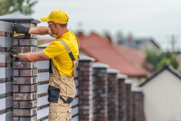 A Fencing Contractor in Galt CA wearing a yellow shirt and overalls working on a brick pillar fence.