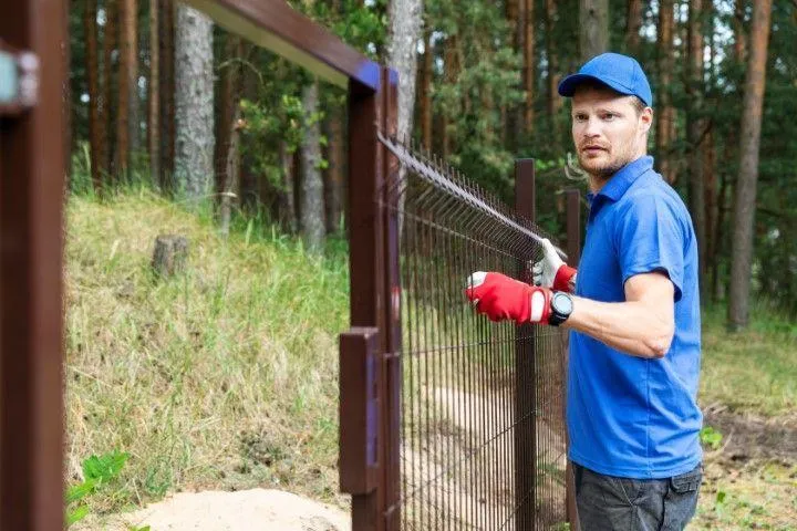 A Fencing Contractor in Galt CA wearing a blue shirt and cap while securing a wire mesh fence.