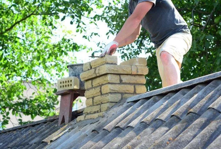 A mason on a corrugated roof carefully repairing a yellow brick chimney, a service from San Gabriel Masonry in San Gabriel CA.