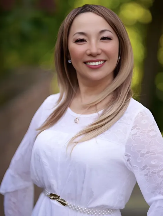 Professional headshot of a smiling Asian woman with blonde-highlighted hair, wearing a white lace top, set against a beautifully blurred outdoor background with green foliage.
