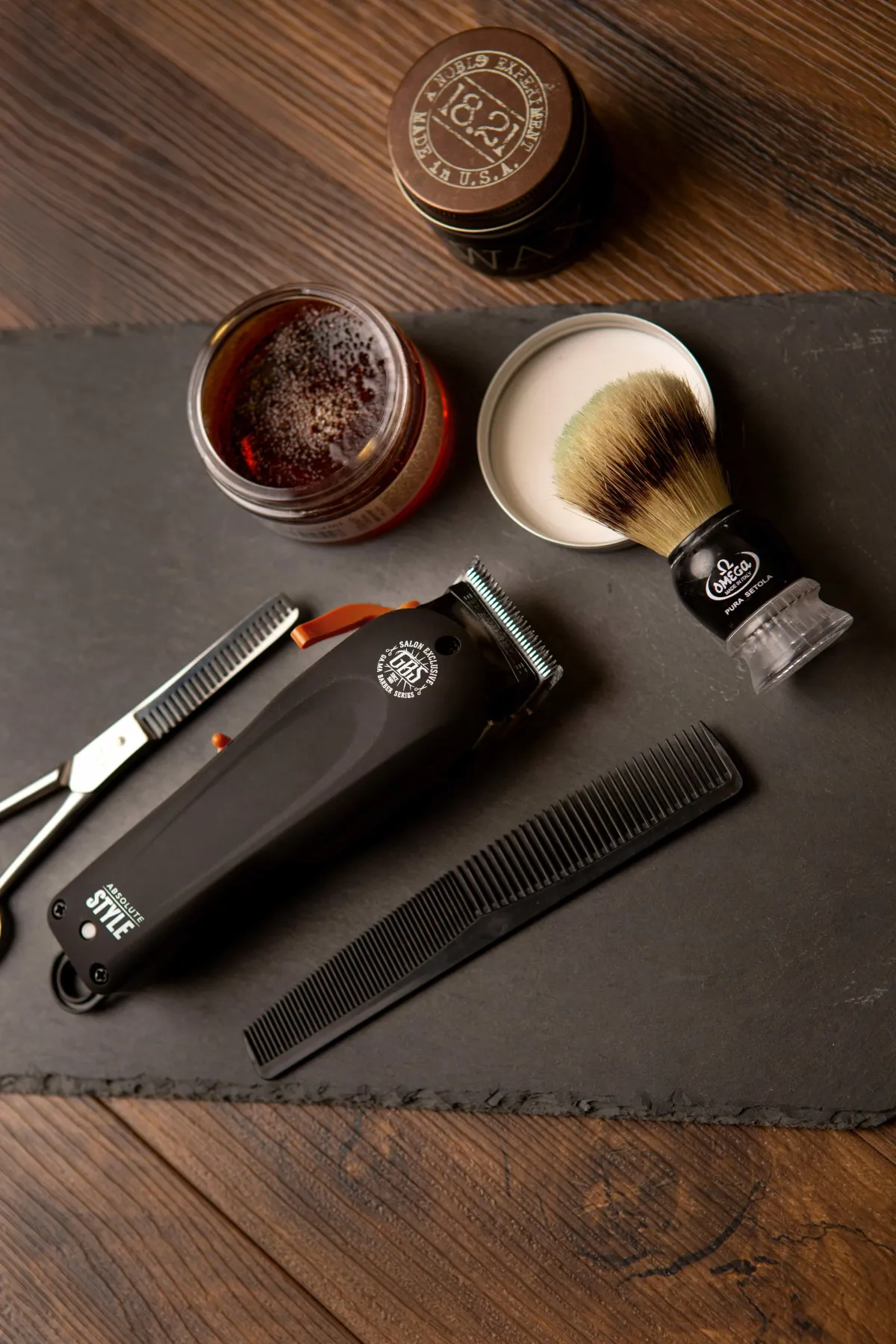 A set of professional barber tools on a slate surface, including clippers, scissors, a comb, a shaving brush, and grooming products, representing essential barbering equipment.