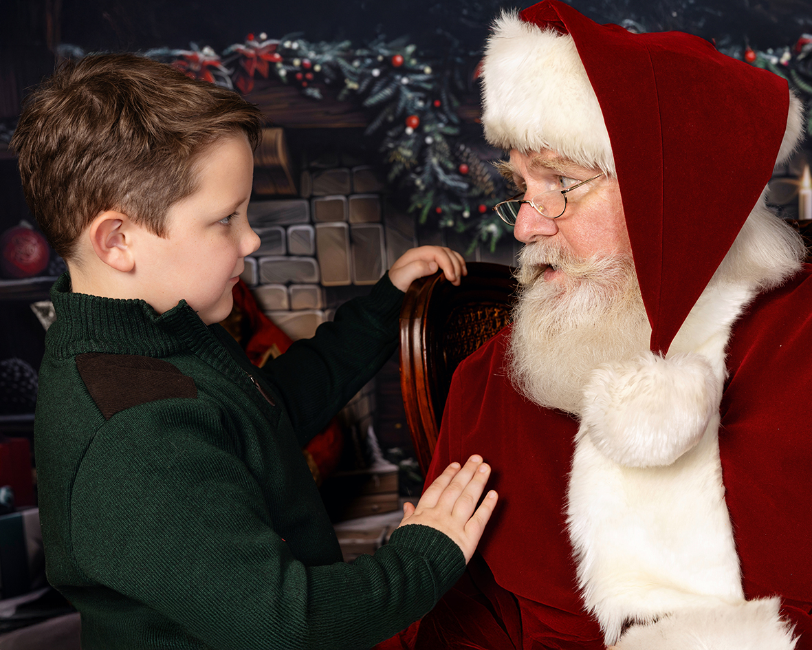 A young boy talks face to face with Santa beside the Christmas fireplace.