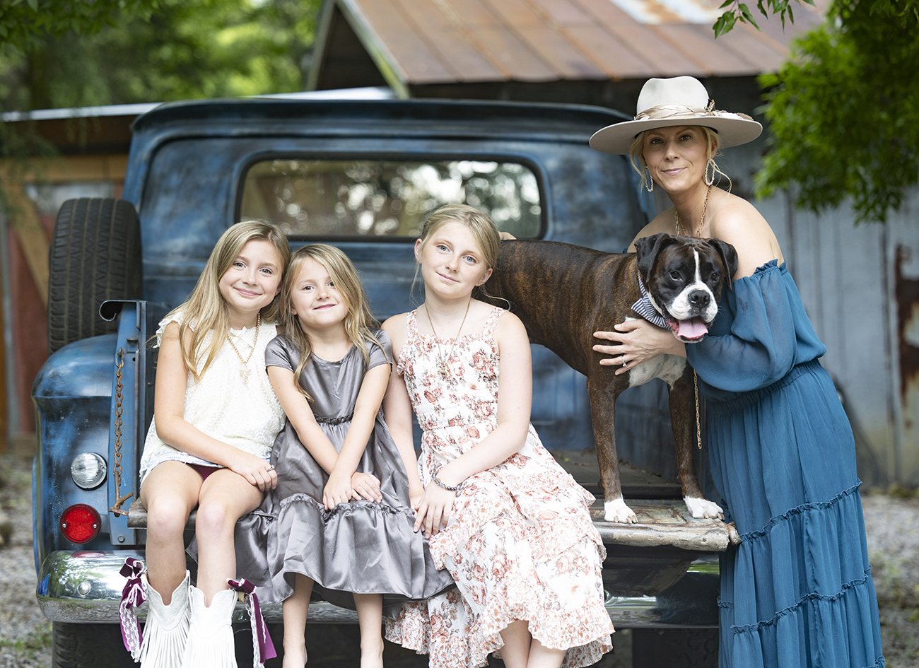 A woman, three girls, and their dog gathered at the back of a vintage truck.