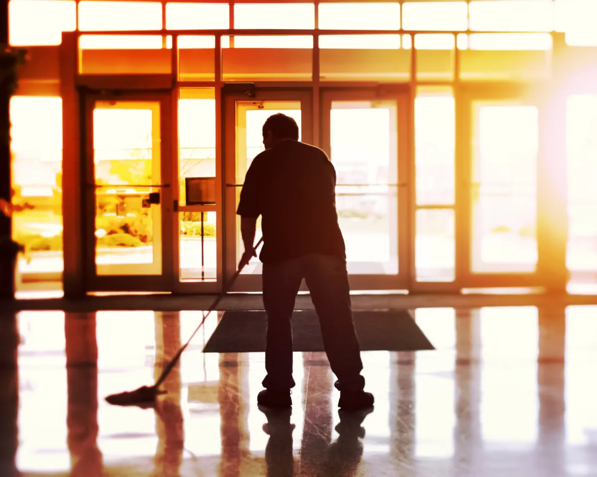 A cleaner works to mop the entryway of a building while the sun shines through the windows . 