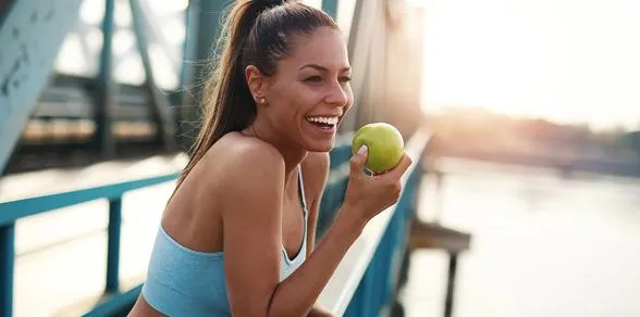 Fit young woman in workout gear smiling and holding a green apple outdoors, symbolizing health, energy, and wellness.
