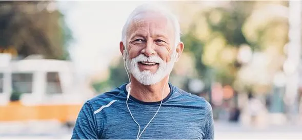 Healthy older man in athletic wear smiling while walking outdoors with earbuds in, reflecting longevity and active aging.