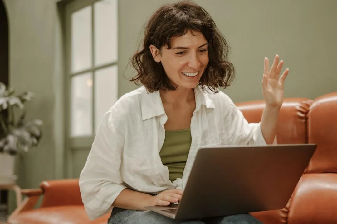 Smiling woman sitting on a sofa, waving at her laptop during a video call in a bright, relaxed home office — representing the confidence and freedom that come from understanding and taking control of your business finances.