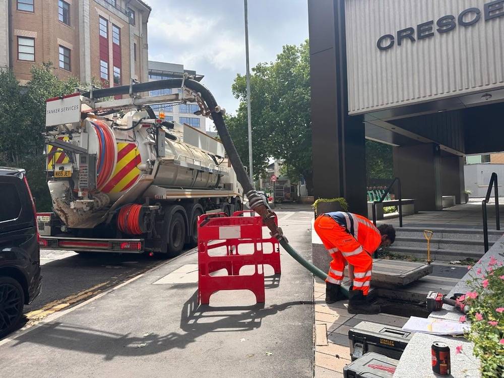 Drainage engineer operating a vacuum tanker for high-pressure drain jetting outside a commercial building