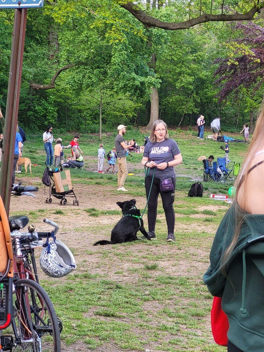 A person in a t-shirt and pants, wearing a treat pouch and holding a leash. A medium black dog sits in front of the person, scratching his chin with his back leg. They stand in the middle of a crowded lawn at a park.