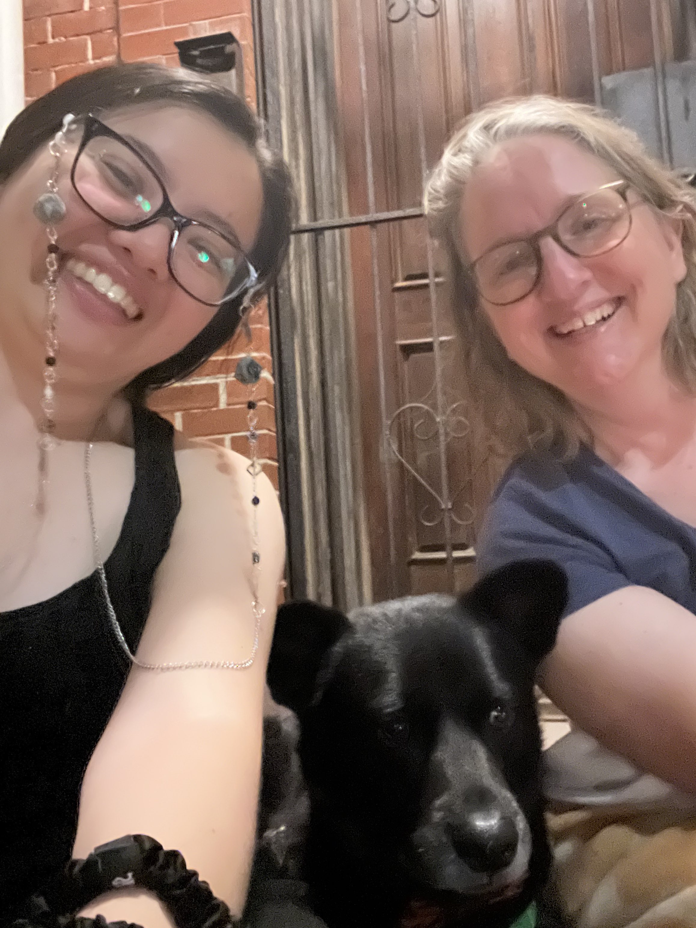 An Asian woman in a black tank top, wearing cats eye glasses holds the selfie camera. A white woman with light, shoulder-length hair leans in on the other side of the image. Between them, a black dog with stand up ears looks off to the side of the camera. 