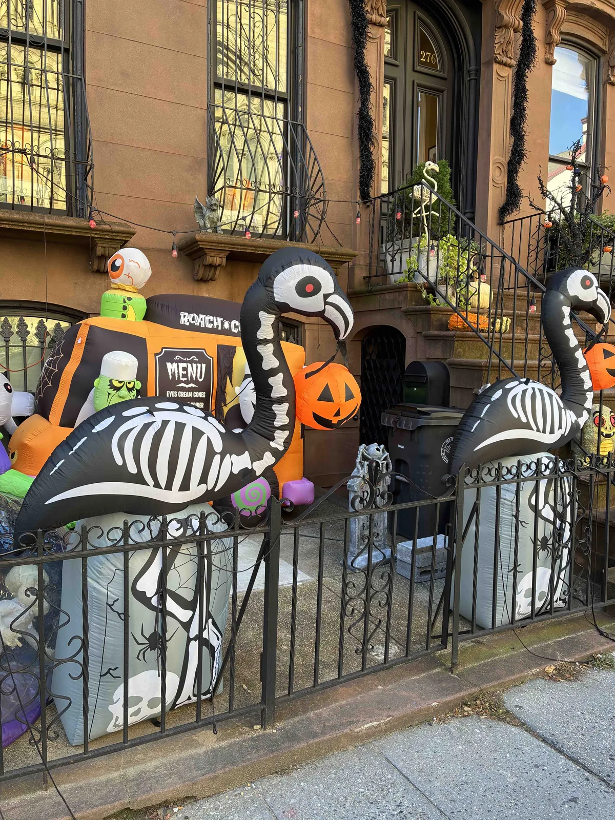 The front yard of a brownstone home. Behind an iron gate are inflatable flamingos as skeletons, a spooky bus, and other Halloween decorations.